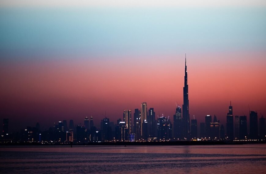 La ligne d'horizon de Dubaï au crépuscule, avec ses bâtiments illuminés, dont le Burj Khalifa, se reflétant sur une eau calme sous un ciel aux nuances changeantes, passant du bleu à l'orange.