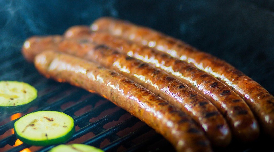 Quatre saucisses grillées et deux tranches de courgette cuisent sur un barbecue, avec des flammes et de la fumée bien visibles.