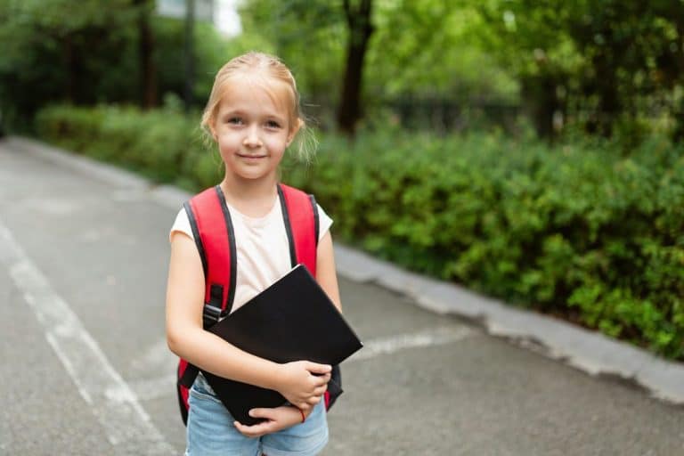 Une jeune fille avec un sac à dos rouge tient un cahier noir alors qu'elle se tient debout sur un chemin entouré de verdure.