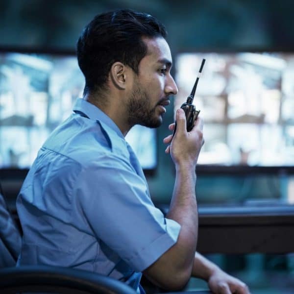 Un agent de sécurité est assis dans une salle de contrôle, parlant dans un talkie walkie, avec plusieurs écrans de surveillance en arrière plan.