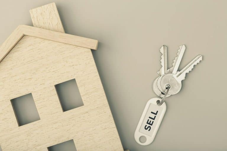Modèle de maison en bois à côté de trois clés sur un porte clés étiqueté « VENDRE » sur fond gris.