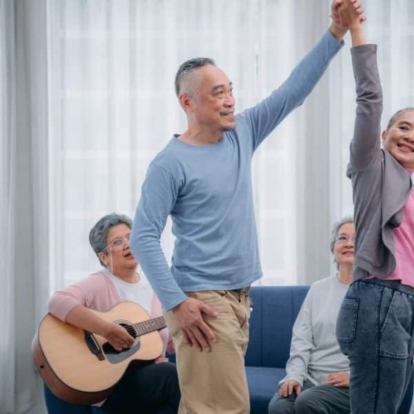 Senior friends are dancing and playing a musical instrument at residence.