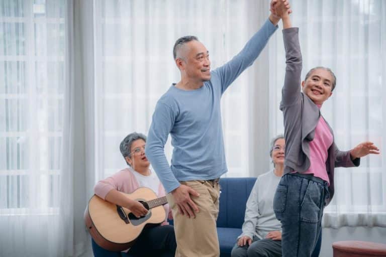 Senior friends are dancing and playing a musical instrument at residence.