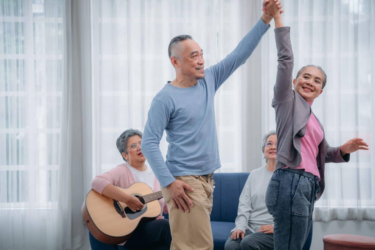 Senior friends are dancing and playing a musical instrument at residence.