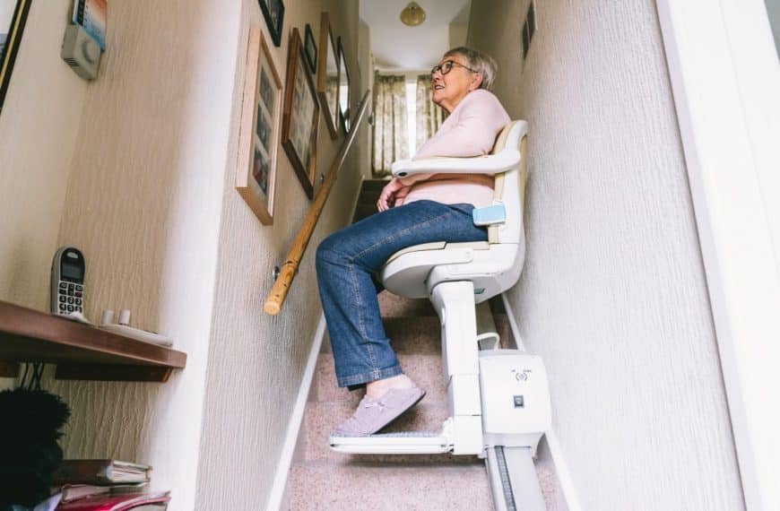 Une femme âgée monte un étroit escalier recouvert de moquette dans un cadre domestique à l'aide d'un monte escalier.