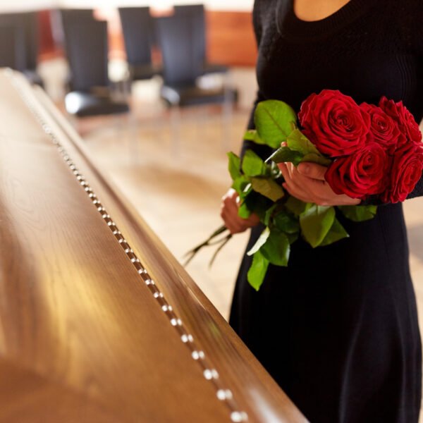 Une personne en robe noire tenant des roses rouges se tient à côté d'un cercueil en bois fermé dans une pièce avec des chaises vides.