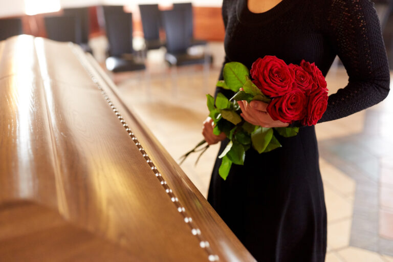 Une personne en robe noire tenant des roses rouges se tient à côté d'un cercueil en bois fermé dans une pièce avec des chaises vides.