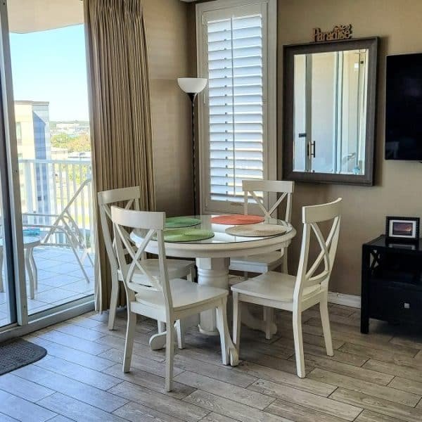 Un coin repas moderne avec une table ronde et quatre chaises. Un balcon avec vue sur la ville est visible à travers des portes coulissantes en verre. Une télévision murale se trouve sur la droite, avec un meuble en dessous.