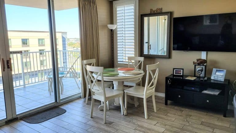 Un coin repas moderne avec une table ronde et quatre chaises. Un balcon avec vue sur la ville est visible à travers des portes coulissantes en verre. Une télévision murale se trouve sur la droite, avec un meuble en dessous.