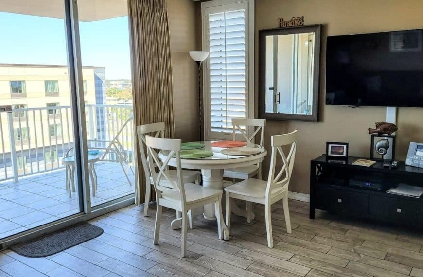 Un coin repas moderne avec une table ronde et quatre chaises. Un balcon avec vue sur la ville est visible à travers des portes coulissantes en verre. Une télévision murale se trouve sur la droite, avec un meuble en dessous.