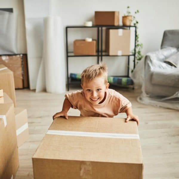 Jeune garçon souriant, appuyé sur un grand carton, dans une pièce remplie de cartons de déménagement et de meubles emballés dans du plastique.