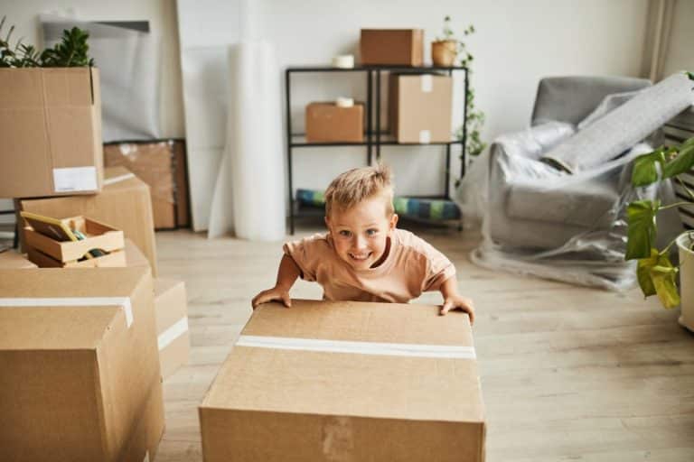 Jeune garçon souriant, appuyé sur un grand carton, dans une pièce remplie de cartons de déménagement et de meubles emballés dans du plastique.