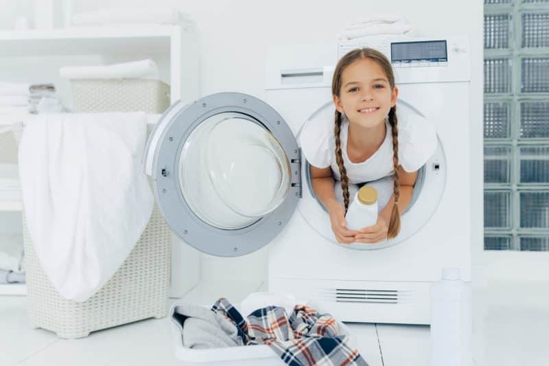 Smiling female child with glad expression, poses in washing machine, holds detergent