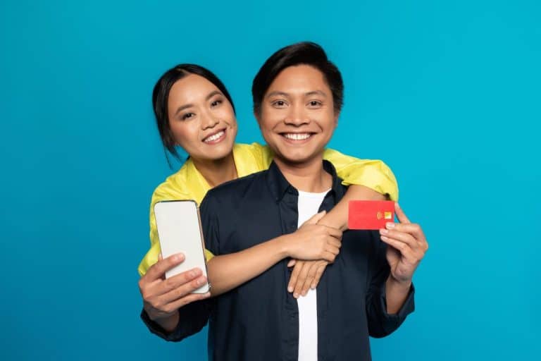 Un couple souriant pose ensemble ; la femme tient un smartphone et l'homme une carte de crédit rouge sur un fond bleu uni.