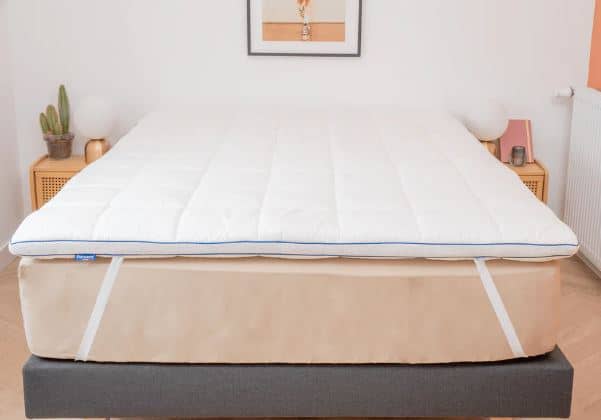 Un lit bien fait avec un surmatelas blanc. La chambre comprend des tables de nuit en bois, des lampes, un cactus et un tableau encadré sur le mur.