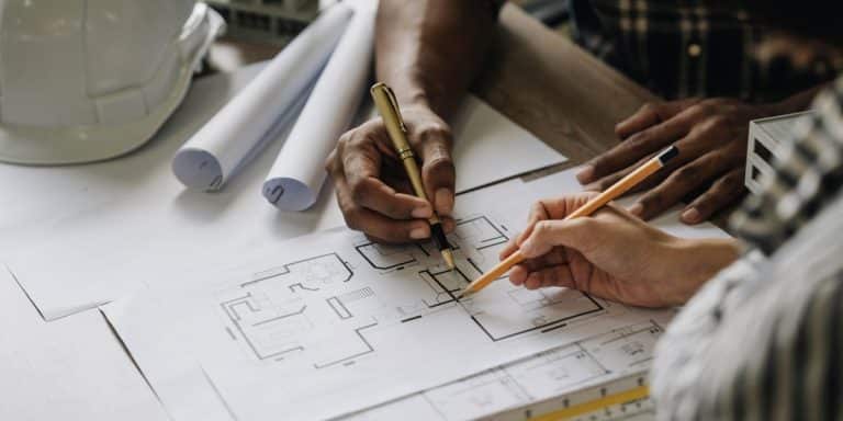 Deux personnes travaillent sur des plans architecturaux à une table, entourées d'un casque, de plans enroulés et d'outils de dessin.