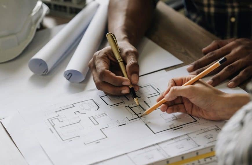 Deux personnes travaillent sur des plans architecturaux à une table, entourées d'un casque, de plans enroulés et d'outils de dessin.