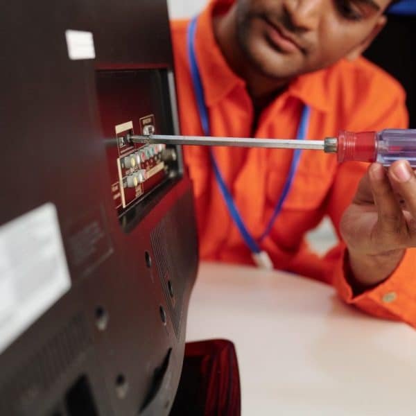 Une personne portant une chemise orange utilise un tournevis pour ajuster ou réparer les ports à l’arrière d’un téléviseur ou d’un moniteur.