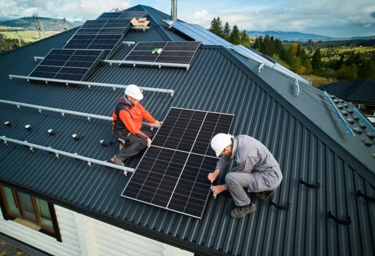 Deux ouvriers portant des casques et des équipements de sécurité installent des panneaux solaires sur un toit en pente sous un ciel partiellement nuageux.