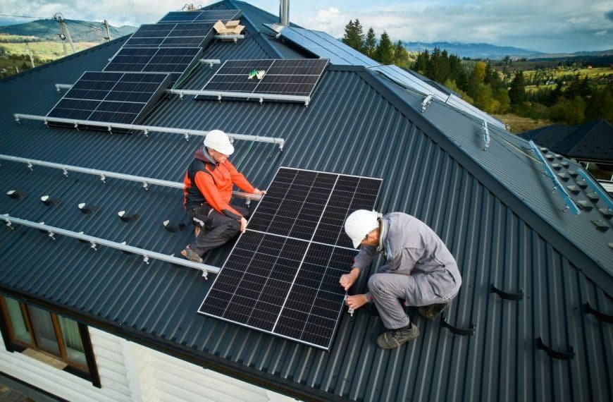 Deux ouvriers portant des casques et des équipements de sécurité installent des panneaux solaires sur un toit en pente sous un ciel partiellement nuageux.
