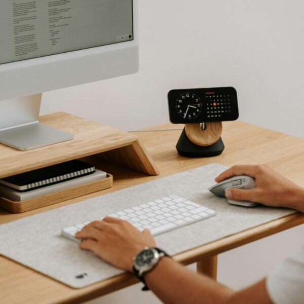 Une personne utilisant une souris et un clavier d'ordinateur sur un bureau en bois bien rangé, avec un écran, une petite horloge, des blocs-notes et des écouteurs.