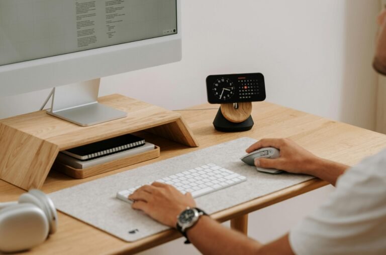 Une personne utilisant une souris et un clavier d'ordinateur sur un bureau en bois bien rangé, avec un écran, une petite horloge, des blocs-notes et des écouteurs.