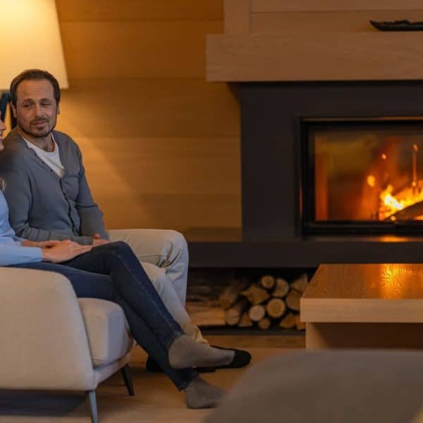 Un homme et une femme sont assis ensemble sur un canapé dans un salon confortable, se tenant par la main et discutant à côté d'une cheminée allumée.