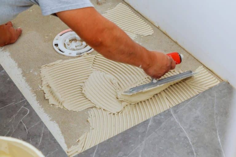 Une personne utilise une truelle pour étaler de la colle à carrelage sur le sol d'une salle de bain près d'un drain, préparant ainsi la surface pour l'installation du carrelage.