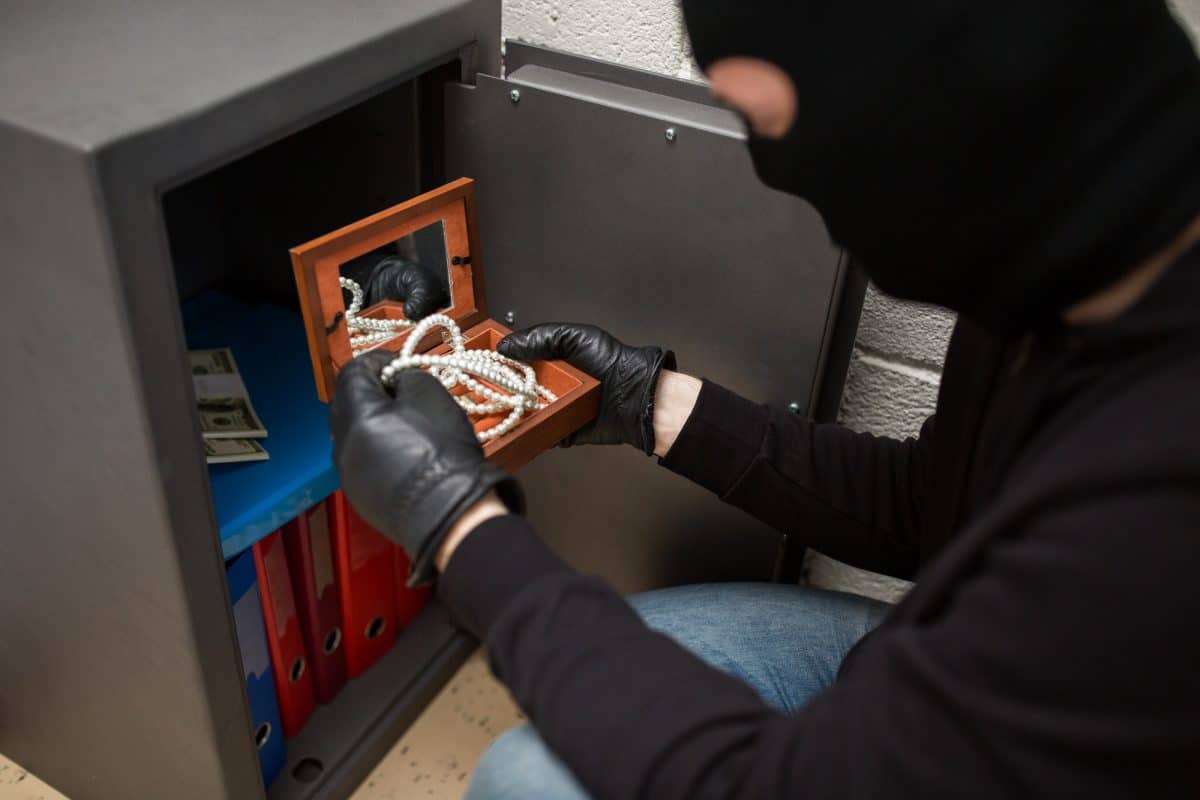 Une personne masquée portant des gants prend une boîte à bijoux remplie de perles et un collier dans un coffre-fort ouvert contenant de l'argent liquide et des dossiers.