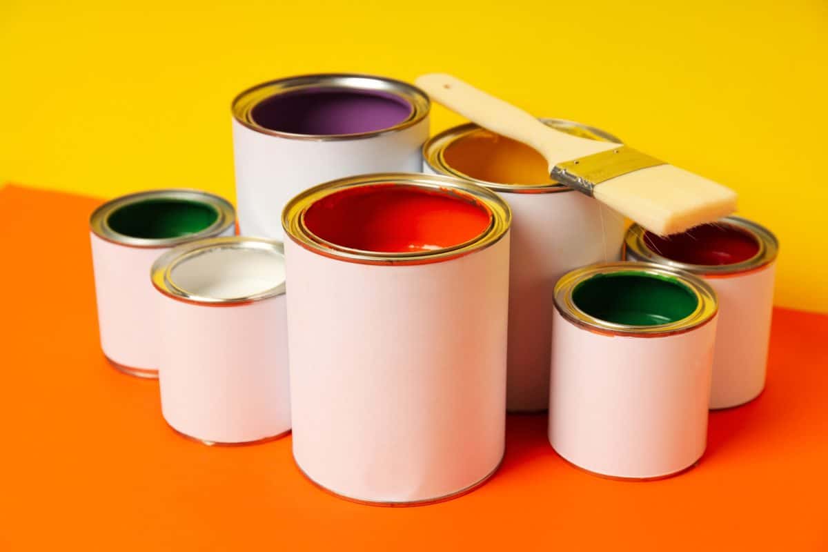 Pots de peinture de différentes couleurs avec un pinceau dessus, disposés sur un fond jaune et orange.