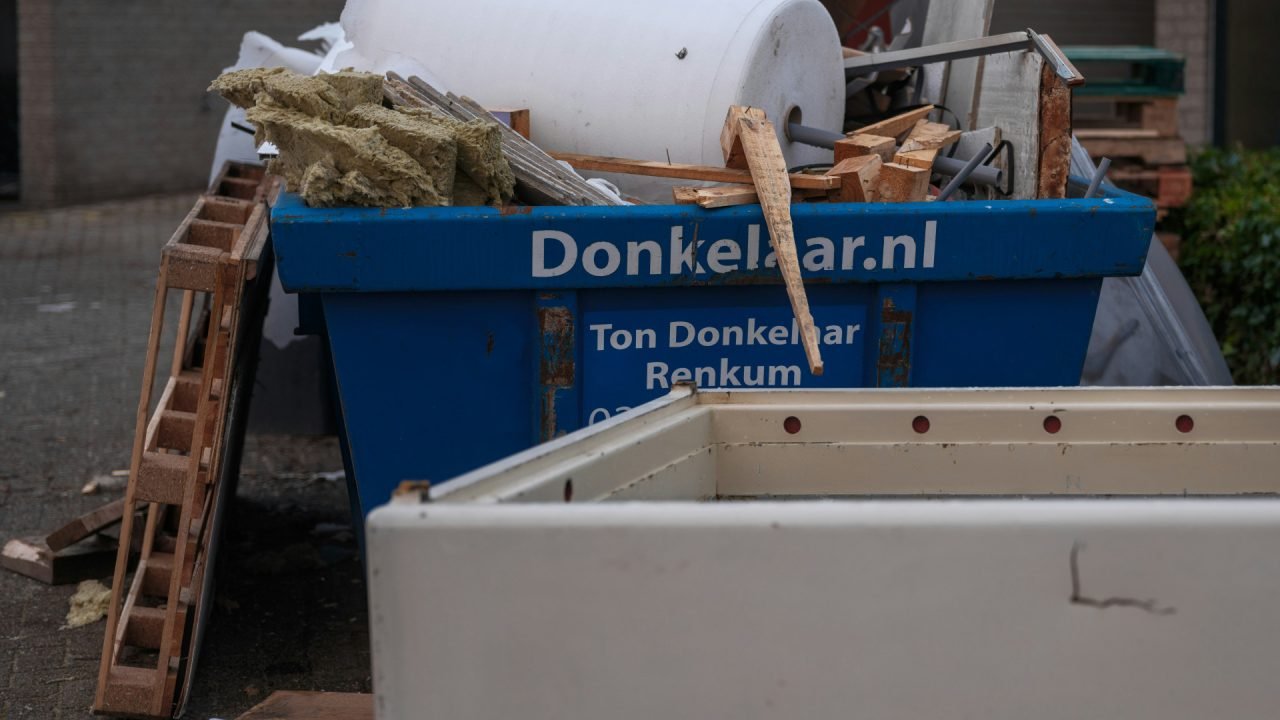Une benne bleue étiquetée "Donkelaar.nl" est remplie de débris de construction, y compris du bois, de l'isolation et des pièces métalliques, avec d'autres débris devant et à côté de la benne.