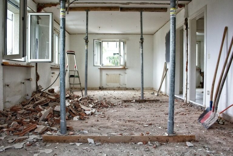 Pièce en cours de rénovation avec des débris sur le sol, des poutres de soutien apparentes, des fenêtres ouvertes, une échelle et des outils de construction visibles.