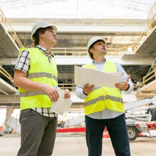 Deux ouvriers du bâtiment portant des gilets de haute visibilité et des casques de sécurité examinent les plans à l'intérieur d'un grand bâtiment en construction.