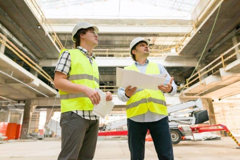 Deux ouvriers du bâtiment portant des gilets de haute visibilité et des casques de sécurité examinent les plans à l'intérieur d'un grand bâtiment en construction.