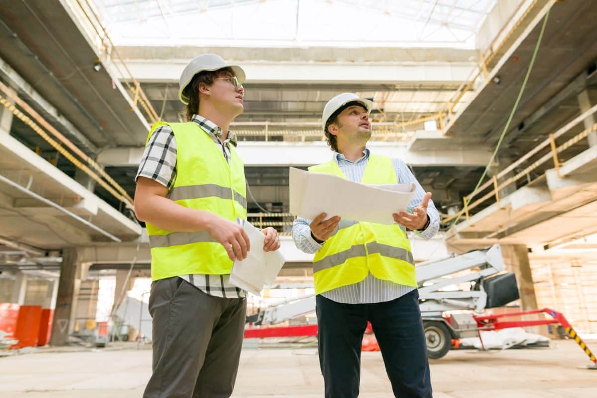 Deux ouvriers du bâtiment portant des gilets de haute visibilité et des casques de sécurité examinent les plans à l'intérieur d'un grand bâtiment en construction.