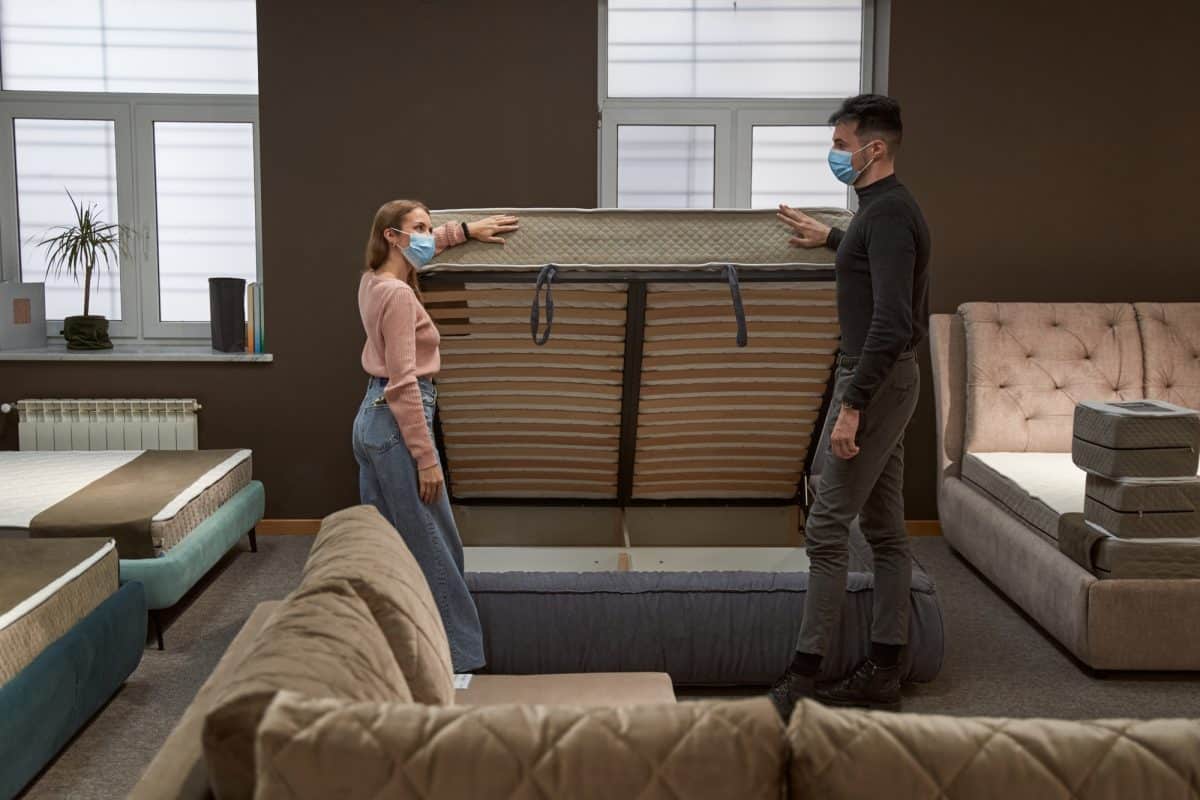 Deux personnes portant des masques examinent un cadre de matelas dans une salle d'exposition avec divers matelas et cadres de lit autour d'eux.