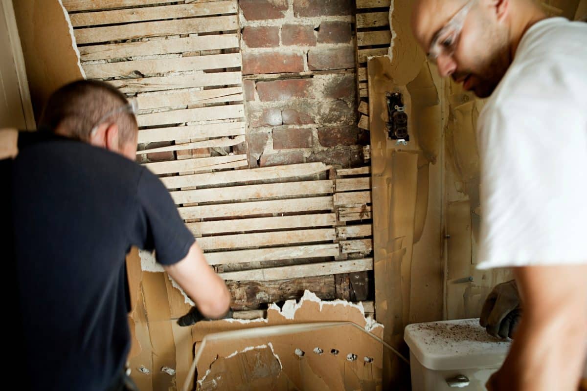 Two men engaged in a home renovation project around a damaged wall.