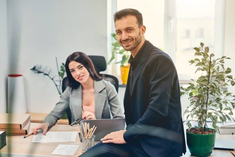 Deux professionnels dans un bureau : une femme en blazer est assise à un bureau avec des documents, et un homme en costume s'appuie sur le bureau en souriant. Des plantes sont visibles en arrière plan.