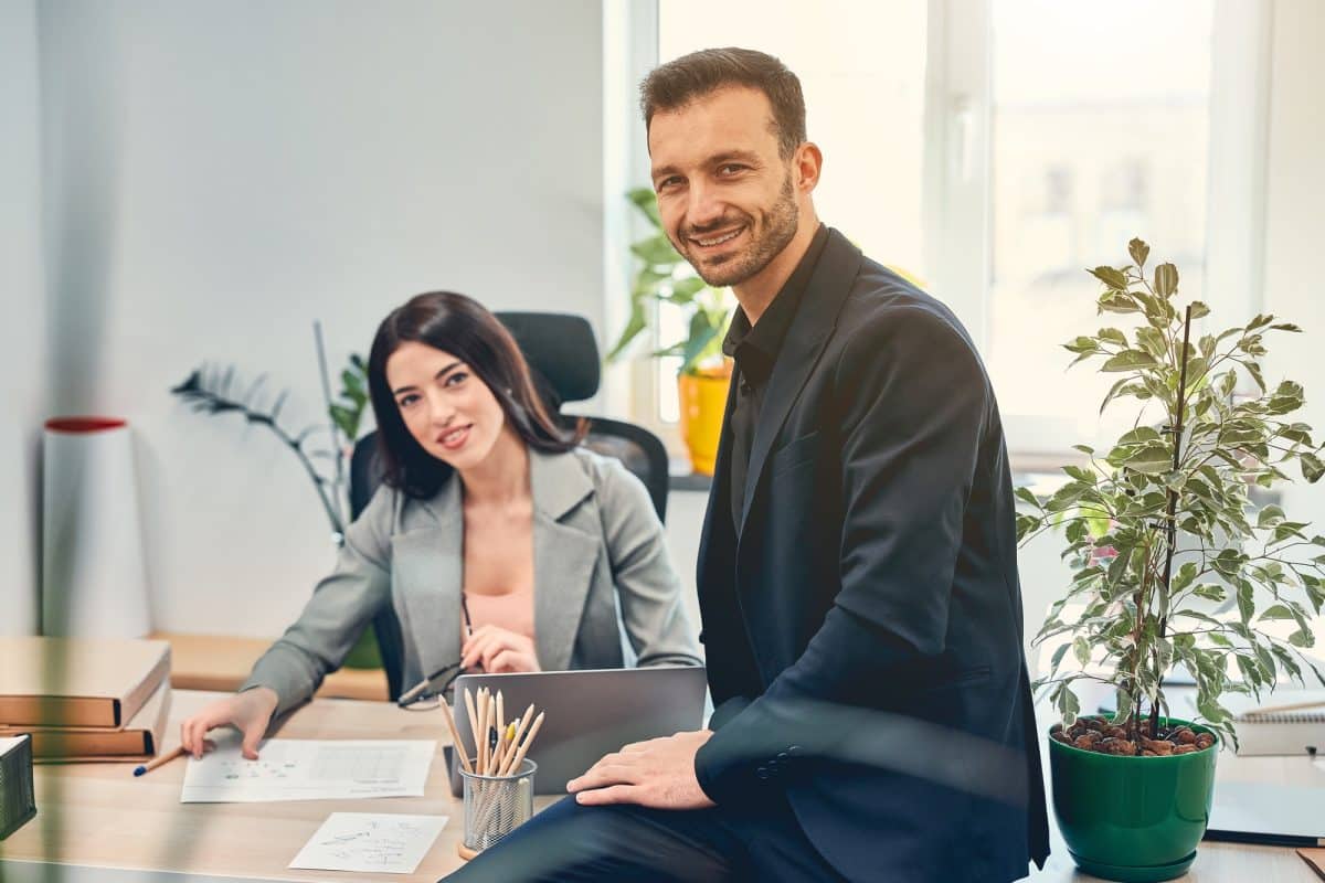 Deux professionnels dans un bureau : une femme en blazer est assise à un bureau avec des documents, et un homme en costume s'appuie sur le bureau en souriant. Des plantes sont visibles en arrière plan.