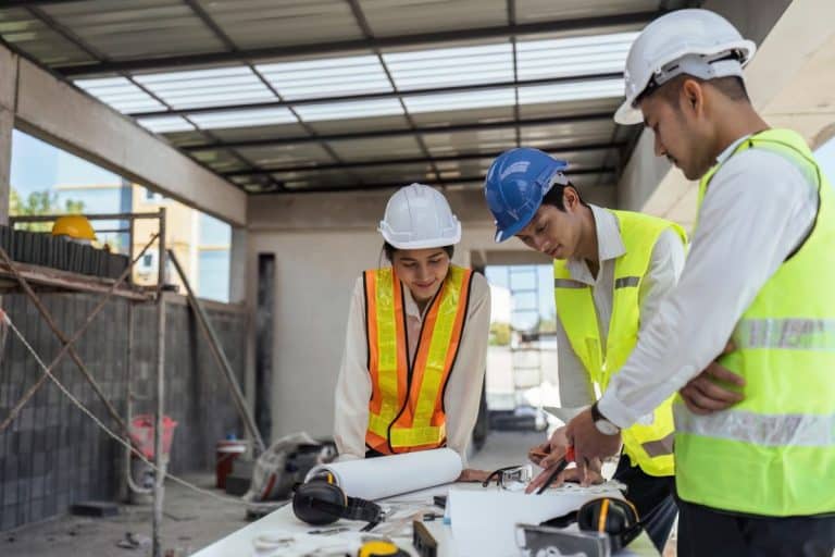 Trois ouvriers du bâtiment portant un équipement de sécurité examinent des plans à une table à l'intérieur d'une structure partiellement construite.