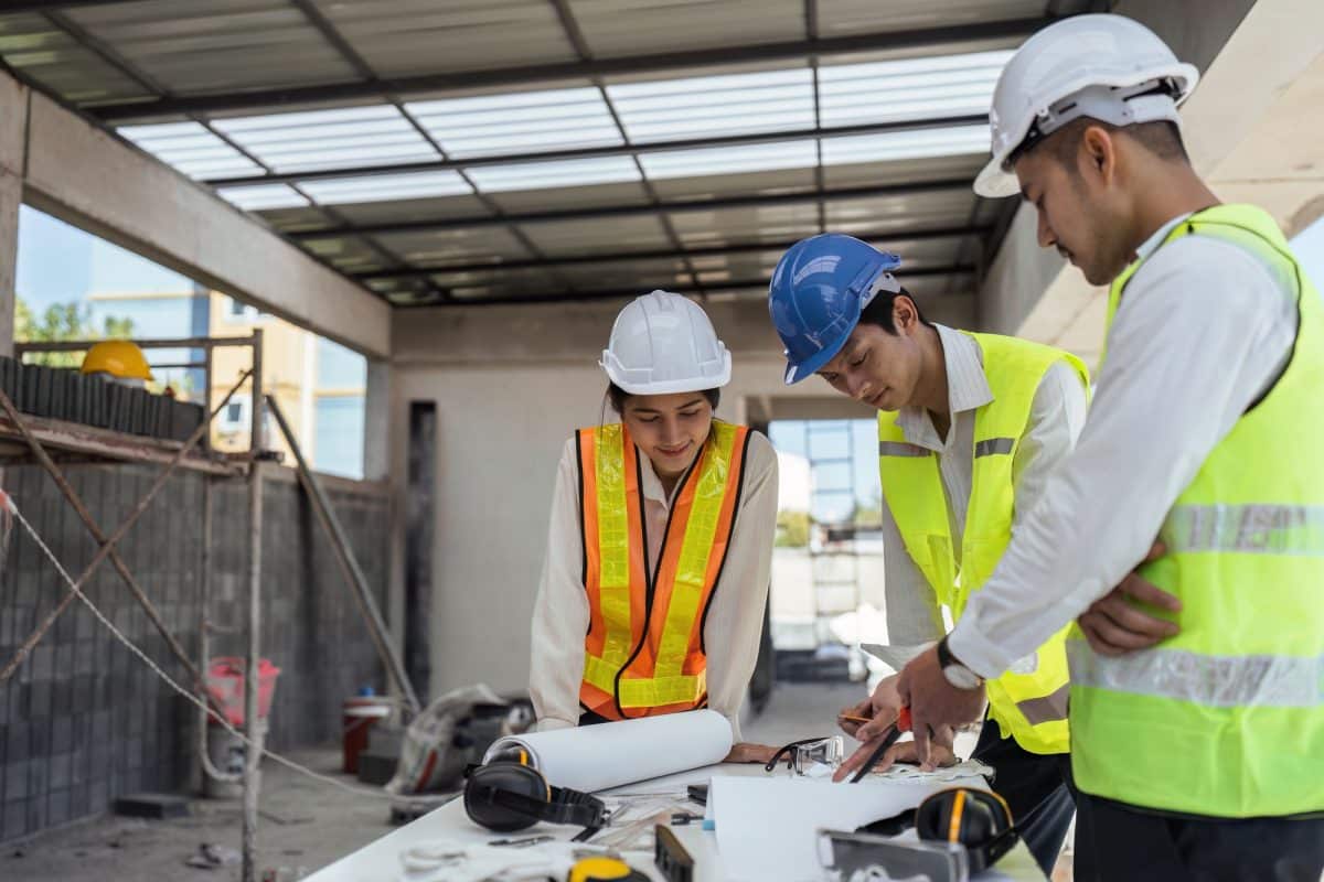 Trois ouvriers du bâtiment portant un équipement de sécurité examinent des plans à une table à l'intérieur d'une structure partiellement construite.