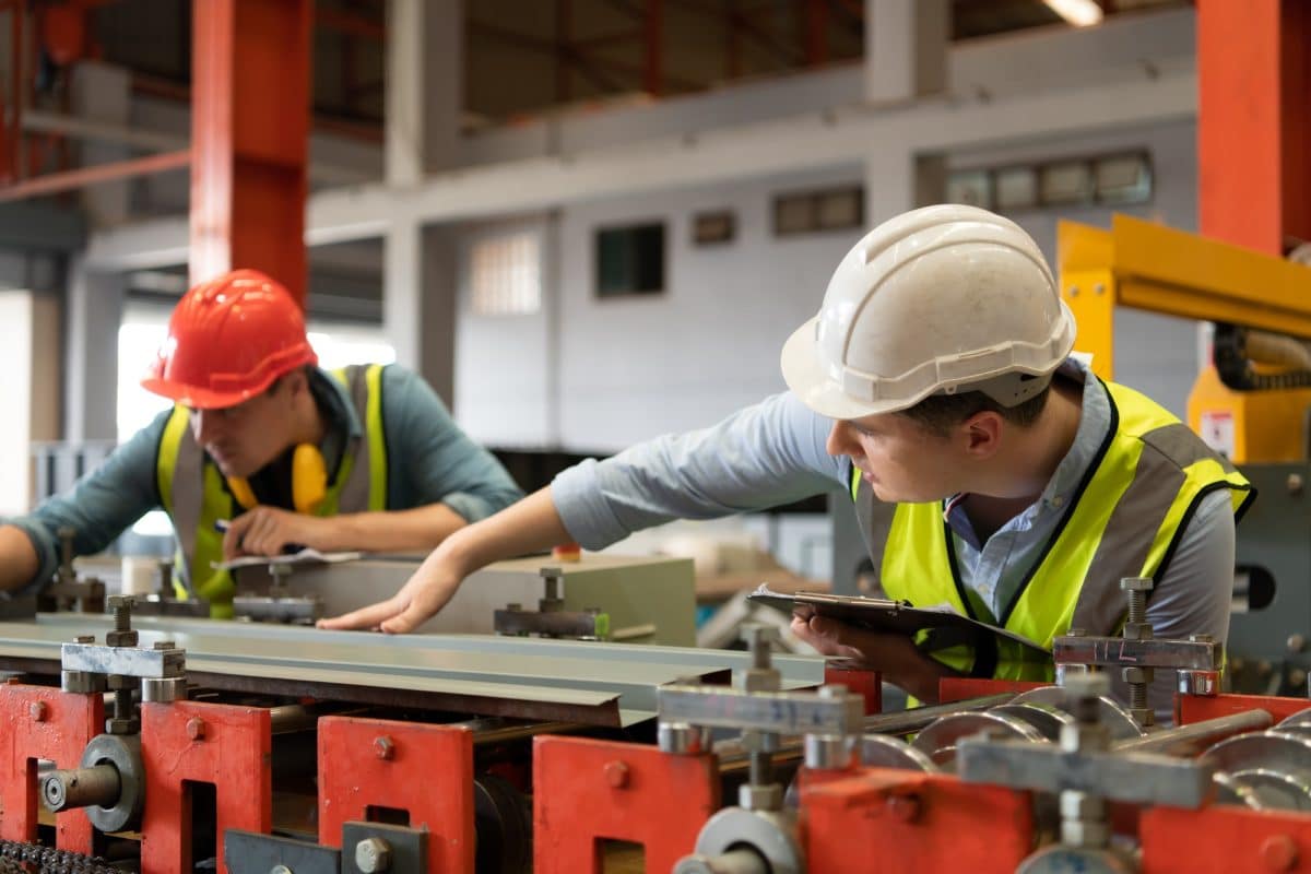 Deux ouvriers portant des casques de sécurité et des gilets de sécurité font fonctionner des machines dans une usine. L'un tient un presse papiers, tandis que l'autre ajuste l'équipement.
