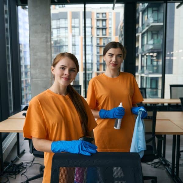 Deux femmes vêtues d'une chemise orange et de gants bleus se tiennent dans un bureau moderne, tenant des produits de nettoyage, avec des bureaux et de grandes fenêtres en arrière plan.