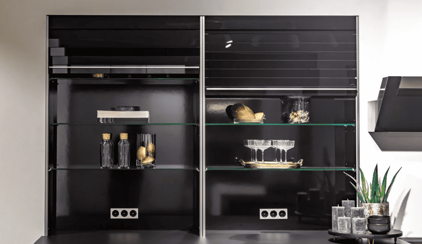 Deux vitrines à étagères en verre, dont l'intérieur est noir, exposent des bouteilles, de la verrerie, des livres et des objets décoratifs. De petites plantes et des tasses sont disposées sur un comptoir situé en dessous. Des prises électriques sont visibles à l'intérieur.