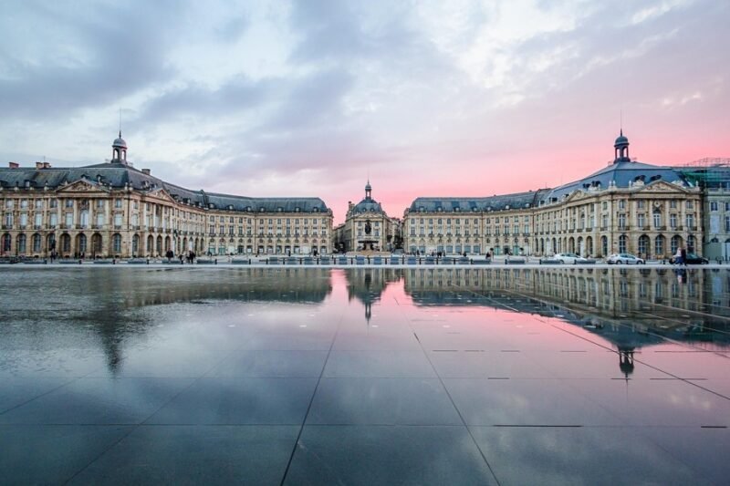 La place de la Bourse à Bordeaux, en France, se reflète dans un grand miroir d'eau au coucher du soleil, sous un ciel partiellement nuageux.