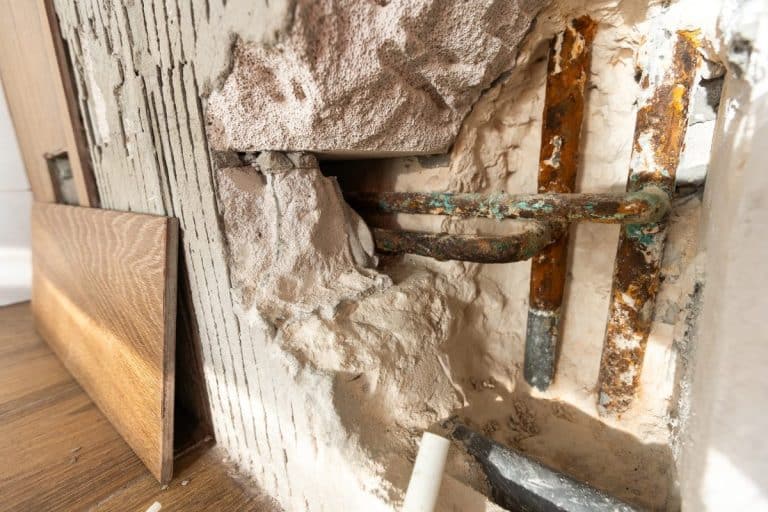 Tuyaux apparents et plâtre endommagé sur un mur, avec une bande de panneaux de bois appuyée à proximité.