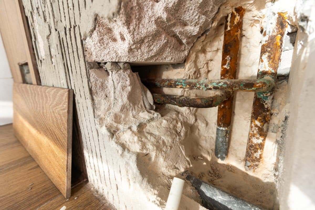 Tuyaux apparents et plâtre endommagé sur un mur, avec une bande de panneaux de bois appuyée à proximité.