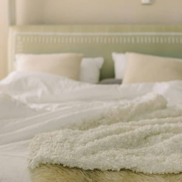 Un lit soigneusement fait avec une tête de lit verte, des draps blancs, des oreillers beiges et une couverture beige texturée pliée au pied. Une lampe est visible sur une table de nuit à côté du lit.