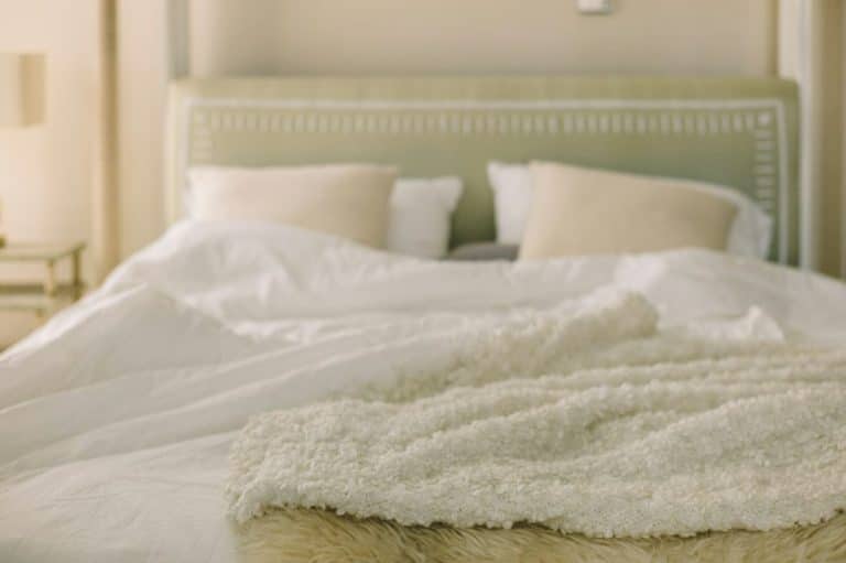 Un lit soigneusement fait avec une tête de lit verte, des draps blancs, des oreillers beiges et une couverture beige texturée pliée au pied. Une lampe est visible sur une table de nuit à côté du lit.