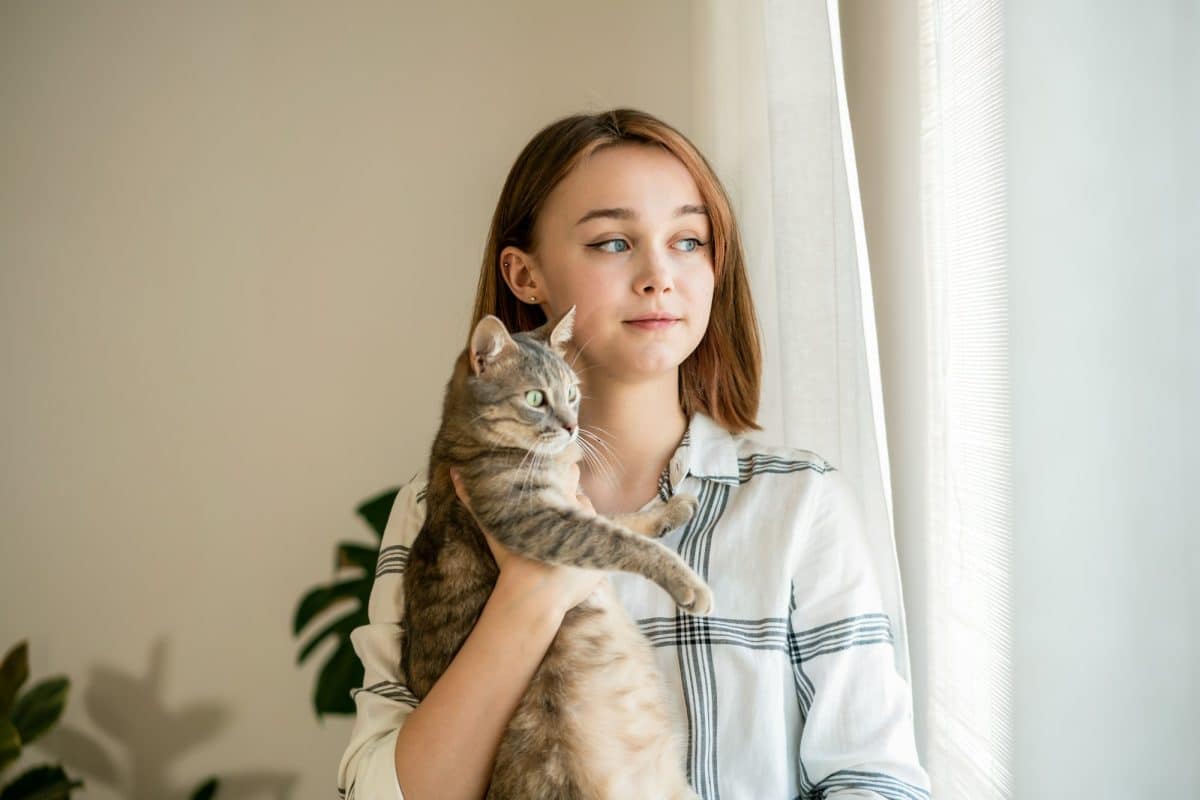 Jeune femme tenant un chat tigré près d'une fenêtre avec des rideaux, toutes deux regardant contemplativement sur le côté.